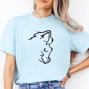 May include: A light blue short-sleeve t-shirt featuring a black line art graphic on the front. The graphic depicts a stylized, abstract silhouette of a person's profile and upper torso. The shirt has a crew neckline and is worn with blue denim jeans.