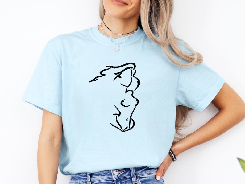 May include: A light blue short-sleeve t-shirt featuring a black line art graphic on the front. The graphic depicts a stylized, abstract silhouette of a person's profile and upper torso. The shirt has a crew neckline and is worn with blue denim jeans.