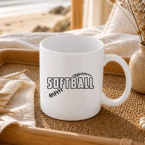 K&ouml;nnte beinhalten: Ein wei&szlig;er Keramikbecher mit einer schwarzen Grafik eines Softballs und dem Wort SOFTBALL, das in fetten, blockartigen Gro&szlig;buchstaben in der Mitte aufgedruckt ist. Der Becher hat einen glatten, zylindrischen K&ouml;rper und einen geschwungenen Henkel.
