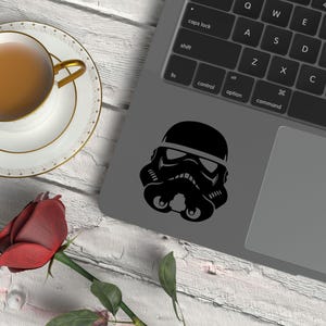 May include: A black Stormtrooper helmet decal applied to the gray surface of a laptop, positioned near a white teacup filled with brown liquid on a saucer and a single red rose with green leaves on a white wooden surface.