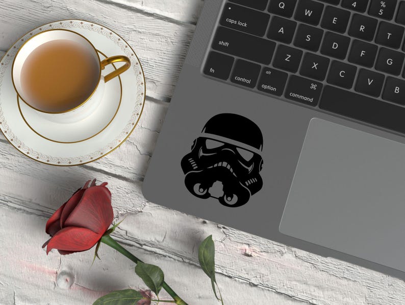 May include: A black Stormtrooper helmet decal applied to the gray surface of a laptop, positioned near a white teacup filled with brown liquid on a saucer and a single red rose with green leaves on a white wooden surface.