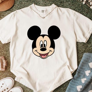 Pu&ograve; includere: Una t-shirt a maniche corte color crema con una grande grafica centrale del volto di Topolino. Il design del personaggio presenta orecchie nere, un viso color marrone chiaro e un'espressione sorridente con una lingua rosa.