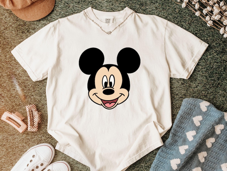 Pu&ograve; includere: Una t-shirt a maniche corte color crema con una grande grafica centrale del volto di Topolino. Il design del personaggio presenta orecchie nere, un viso color marrone chiaro e un'espressione sorridente con una lingua rosa.