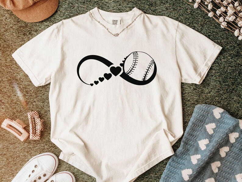 K&ouml;nnte beinhalten: Ein cremefarbenes Kurzarm-T-Shirt mit einem schwarzen Grafikdruck eines Unendlichkeitssymbols. Das Symbol enth&auml;lt eine Reihe kleiner Herzen, die an Gr&ouml;&szlig;e zunehmen und in eine Baseball-Illustration auf der rechten Seite &uuml;bergehen.