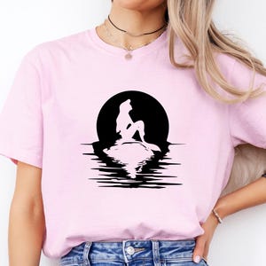 May include: A light pink short-sleeve t-shirt featuring a black graphic print of a mermaid silhouette sitting on a rock against a large circular moon, with the reflection of the mermaid and the moon visible in the water below the rock.