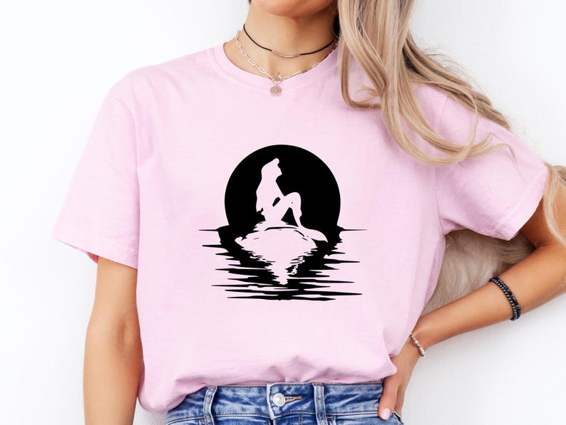 May include: A light pink short-sleeve t-shirt featuring a black graphic print of a mermaid silhouette sitting on a rock against a large circular moon, with the reflection of the mermaid and the moon visible in the water below the rock.
