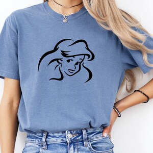 May include: A blue short-sleeve crew neck t-shirt featuring a black line art graphic of a female character with long flowing hair centered on the chest. The shirt is made of a soft, washed-out fabric and is paired with blue denim jeans.