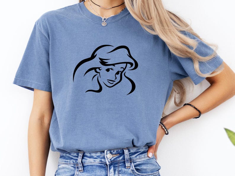 May include: A blue short-sleeve crew neck t-shirt featuring a black line art graphic of a female character with long flowing hair centered on the chest. The shirt is made of a soft, washed-out fabric and is paired with blue denim jeans.