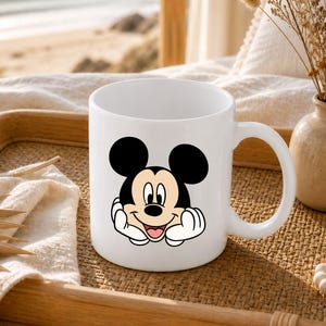 Pu&ograve; includere: Una tazza in ceramica bianca con un'illustrazione grafica di Topolino che appoggia il mento sulle mani. Il personaggio ha orecchie nere, un viso color carne e un ampio sorriso. La tazza ha un corpo cilindrico liscio con un manico curvo laterale.