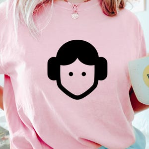 May include: A light pink short-sleeved t-shirt featuring a black minimalist graphic of a stylized face with round hair buns. The shirt is worn with a gold necklace and is held next to a light blue mug with a yellow smiley face design.