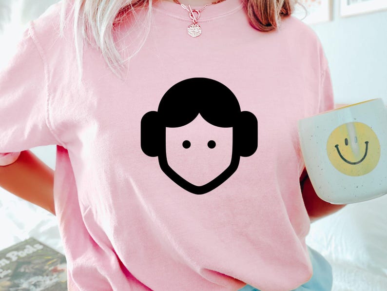 May include: A light pink short-sleeved t-shirt featuring a black minimalist graphic of a stylized face with round hair buns. The shirt is worn with a gold necklace and is held next to a light blue mug with a yellow smiley face design.