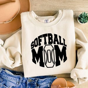 K&ouml;nnte beinhalten: Ein cremefarbenes Sweatshirt mit Rundhalsausschnitt und dem schwarzen Schriftzug SOFTBALL MOM auf der Vorderseite, wobei der Buchstabe O als Softball mit sichtbaren roten N&auml;hten gestaltet ist. Das Kleidungsst&uuml;ck hat lange &Auml;rmel sowie gerippte B&uuml;ndchen.