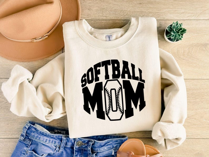 K&ouml;nnte beinhalten: Ein cremefarbenes Sweatshirt mit Rundhalsausschnitt und dem schwarzen Schriftzug SOFTBALL MOM auf der Vorderseite, wobei der Buchstabe O als Softball mit sichtbaren roten N&auml;hten gestaltet ist. Das Kleidungsst&uuml;ck hat lange &Auml;rmel sowie gerippte B&uuml;ndchen.