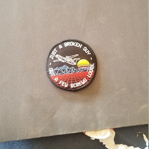 May include: A circular embroidered patch with a black border featuring a white wireframe airplane, a yellow sun, and a red grid landscape. The text reads JUST A BROKEN GUY - GOT A FEW SCREWS LOOSE in white lettering around the circular edge.