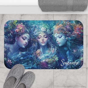 May include: A rectangular bath mat featuring a fantasy illustration of three mermaids with floral headdresses and flowing hair. The mermaids are surrounded by aquatic plants and marine life, with the name "Suzanne" in cursive.