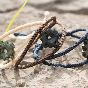 May include: Three braided rope bracelets with green jade stone charms. The charms are shaped like a star with a hole in the center. The bracelets are in different colors: beige, brown, and navy blue.