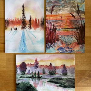 Puede incluir: Tres pinturas de acuarela de paisajes. Una representa una escena nevada con árboles altos y un arroyo azul. Otra muestra una puesta de sol sobre el agua con hierbas altas. La tercera pintura muestra una escena de bosque brumoso con un lago reflectante.