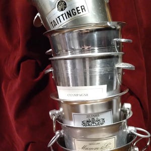 Könnte beinhalten: Ein Stapel silberner Champagnerkühler mit verschiedenen Markenetiketten, darunter "Taittinger" und "Canard-Duchêne". Die Eimer haben Griffe und sind vor einem dunkelroten Hintergrund angeordnet.