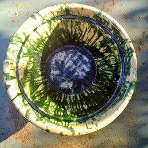 Puede incluir: Un cuenco de cerámica hecho a mano con un diseño único. El cuenco presenta una vibrante combinación de colores verde y azul con un patrón circular. El cuenco está sobre una superficie azul claro.