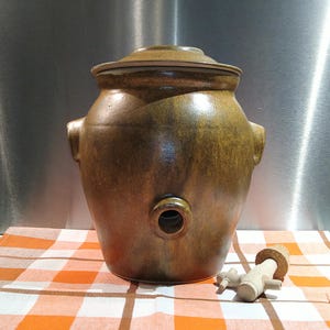 May include: A large, brown ceramic fermentation crock with a lid, two side handles, and a spigot. A wooden spigot is placed next to the crock on a checkered orange and white cloth. The crock has a rustic, glazed finish.