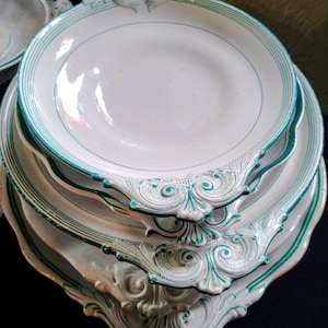 Könnte beinhalten: Ein Stapel weißer Keramikteller mit einem türkis-weißen Zierrand. Die Teller haben einen gewellten Rand mit verzierten türkisfarbenen Akzenten. Die Teller sind übereinander gestapelt.