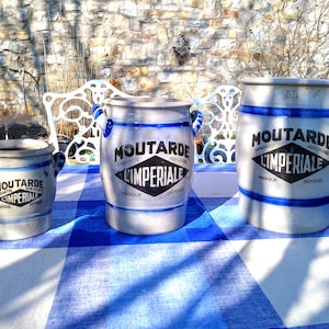 Puede incluir: Tres tarros de mostaza de cerámica vintage con detalles azules y asas. Los tarros están etiquetados como "Moutarde Imperiale" en texto negro. Están sobre un mantel a cuadros azul y blanco, con una pared de piedra y un banco blanco al fondo.