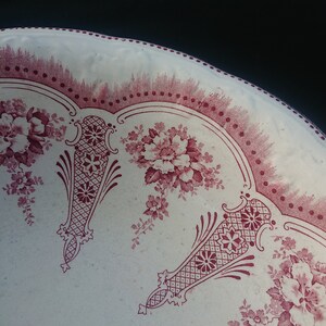 May include: Close-up of a white plate with a burgundy floral pattern. The plate features a repeating design of flowers and decorative elements. The rim has a dotted border. The plate is likely vintage or antique.