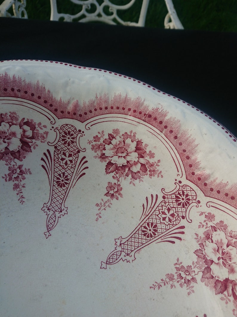 May include: Close-up of a white plate with a burgundy floral pattern. The plate features a repeating design of flowers and decorative elements. The rim has a dotted border. The plate is likely vintage or antique.