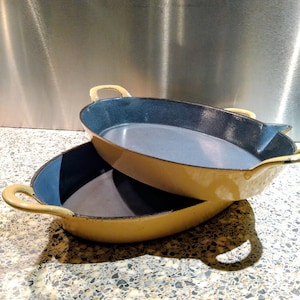 May include: Two yellow oval cast iron pans with blue interiors are stacked. Each pan has a handle on either side. The pans are on a speckled surface with a stainless steel background.