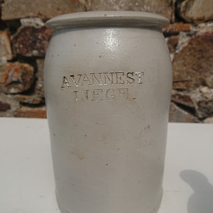 Puede incluir: Un tarro de gres gris claro con tapa. El tarro tiene el texto en relieve "AVANNEST LIEGE". El tarro es cil&iacute;ndrico con una base ligeramente m&aacute;s ancha y una parte superior redondeada. El fondo es una pared de piedra.