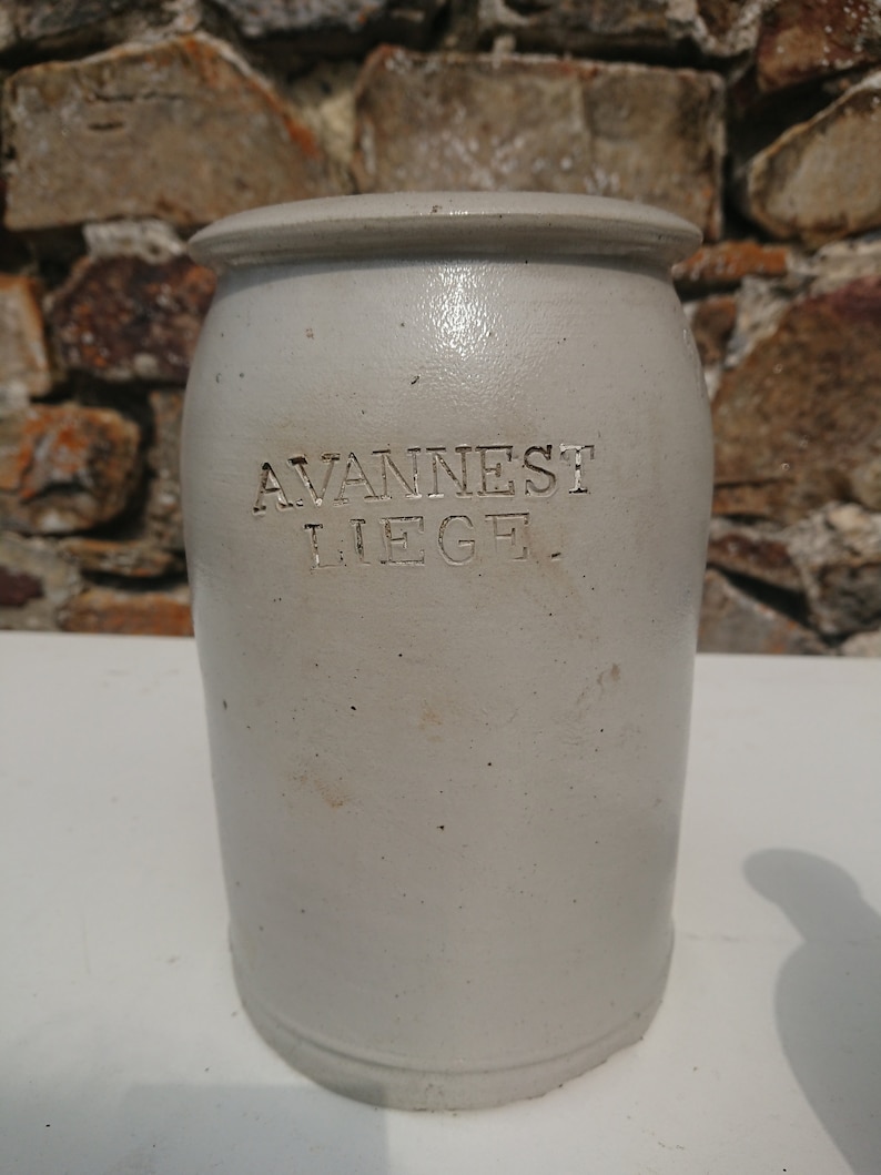 Puede incluir: Un tarro de gres gris claro con tapa. El tarro tiene el texto en relieve "AVANNEST LIEGE". El tarro es cil&iacute;ndrico con una base ligeramente m&aacute;s ancha y una parte superior redondeada. El fondo es una pared de piedra.