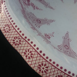 May include: Close-up of a white ceramic plate with a burgundy floral pattern. The plate's rim features a repeating geometric design with flower accents and a dotted border. The plate's interior has a detailed floral design.