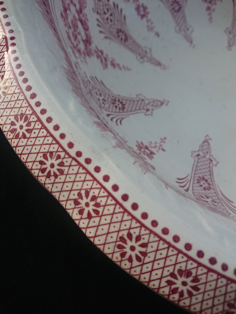 May include: Close-up of a white ceramic plate with a burgundy floral pattern. The plate's rim features a repeating geometric design with flower accents and a dotted border. The plate's interior has a detailed floral design.