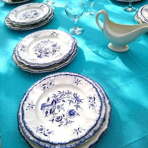 May include: A collection of white porcelain plates with blue floral patterns and scalloped edges. The plates are stacked on a turquoise tablecloth, alongside wine glasses and a gravy boat. The design features birds and botanical elements.