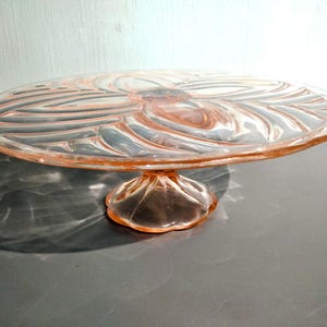 Puede incluir: Un soporte para pasteles de cristal rosa vintage. El soporte presenta una base redonda y festoneada que sostiene un plato circular con un diseño detallado y arremolinado. El cristal tiene un ligero tono rosado, ideal para pasteles.