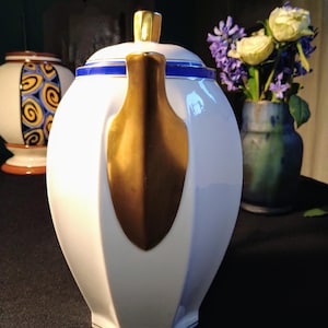 Puede incluir: Tetera de porcelana blanca con asa dorada y ribete azul. La tetera tiene una forma geométrica y una tapa con un pomo dorado.