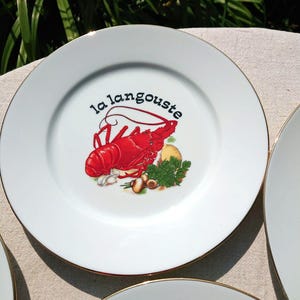 Puede incluir: Plato de cerámica blanco con borde dorado, con una ilustración de langosta roja y las palabras "la langouste" en escritura negra. El plato también incluye ilustraciones de verduras. Se ve un juego de platos.