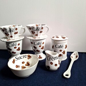 Peut inclure: Un ensemble à café en céramique blanche comprenant deux filtres à café cannelés, deux tasses assorties, un pot avec couvercle, un petit pot à crème, un sucrier et une cuillère. Chaque pièce est ornée de grains de café et de texte noir.