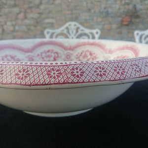 May include: A white ceramic bowl with a decorative red floral pattern around the rim and interior. The rim features a repeating geometric design with small flower motifs. The bowl is set against a blurred background.