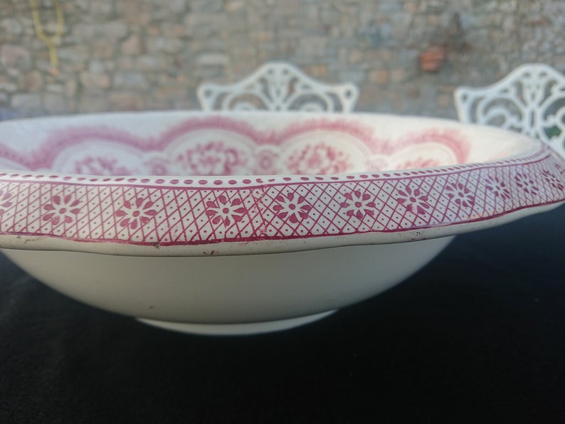 May include: A white ceramic bowl with a decorative red floral pattern around the rim and interior. The rim features a repeating geometric design with small flower motifs. The bowl is set against a blurred background.