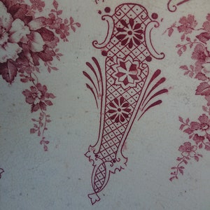 May include: Close-up of a white surface with a burgundy floral and geometric pattern. The design includes stylized flowers, vines, and a central decorative element with a diamond grid and floral accents. The overall aesthetic is vintage.
