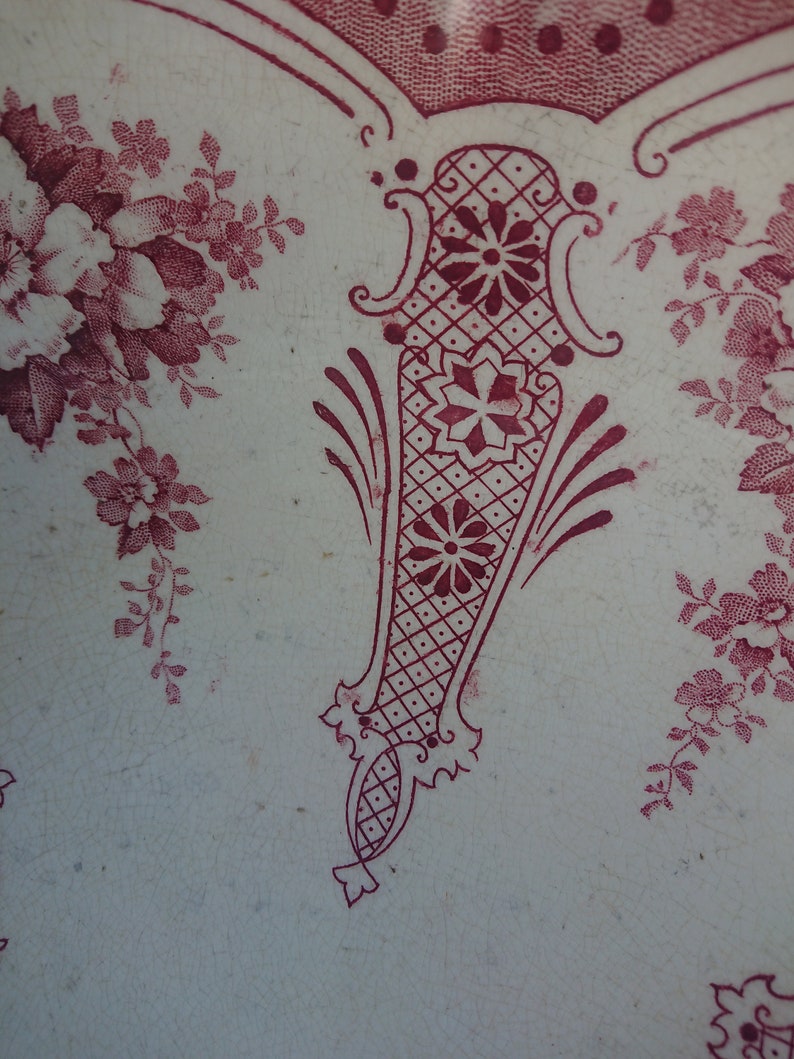 May include: Close-up of a white surface with a burgundy floral and geometric pattern. The design includes stylized flowers, vines, and a central decorative element with a diamond grid and floral accents. The overall aesthetic is vintage.
