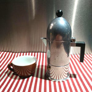 May include: A silver-coloured metal stovetop espresso maker with a black handle and a black knob on the lid, positioned next to a brown ceramic cup with a white interior. The espresso maker features a cylindrical body and a small brass-coloured valve.
