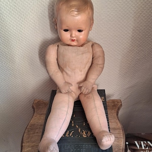 May include: Vintage baby doll with a painted face, seated on a dark book. The doll has a molded head, painted features, and a cloth body. The doll's arms and legs are made of a soft material. The doll is a classic toy.