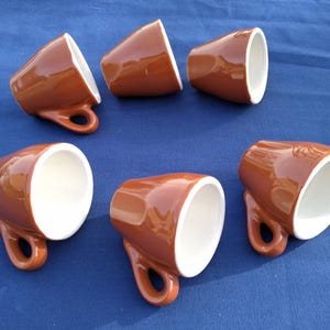 Könnte beinhalten: Sechs braune Espresso-Tassen aus Keramik mit weißem Innenraum sind auf einer blauen Oberfläche angeordnet. Jede Tasse hat einen abgerundeten Henkel und eine glänzende Oberfläche. Die Tassen sind angewinkelt, um ihr Inneres zu zeigen.