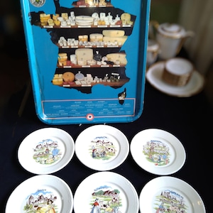 Könnte beinhalten: Sechs weiße Keramikteller mit bunten Illustrationen von französischen Landszenen und Käse. Jeder Teller zeigt eine andere Region Frankreichs, darunter die Normandie, Savoyen und die Auvergne. Die Teller sind mit einem blauen Rand und den Worten "Fromages de France" verziert.