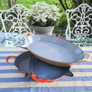 May include: Two oval-shaped, enamelware pans in the image. One is light brown with a light blue interior and tan handles. The other is dark blue with red handles. A silver spoon rests on a blue and white striped tablecloth.