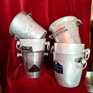 Op de afbeelding: Een verzameling van vier zilverkleurige metalen champagnekoelers, elk voorzien van verschillende merklabels, waaronder Champagne Lamotte & Co, Champagne Gauthier en andere vintage logo's. De emmers zijn voorzien van handgrepen aan de zijkant.