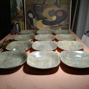 Könnte beinhalten: Ein Set aus zwölf rechteckigen Keramikplatten. Jede Platte hat ein strukturiertes Muster in Blau-, Grün- und Goldtönen mit einem Goldrand. Die Teller sind auf einer rosa Tischdecke angeordnet, was auf einen Ess- oder Servierbereich hindeutet.