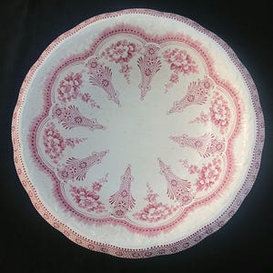 May include: A round, white ceramic serving platter with a scalloped edge. The platter features a pink floral and geometric design around the rim and center. The floral pattern includes detailed flowers and decorative elements.